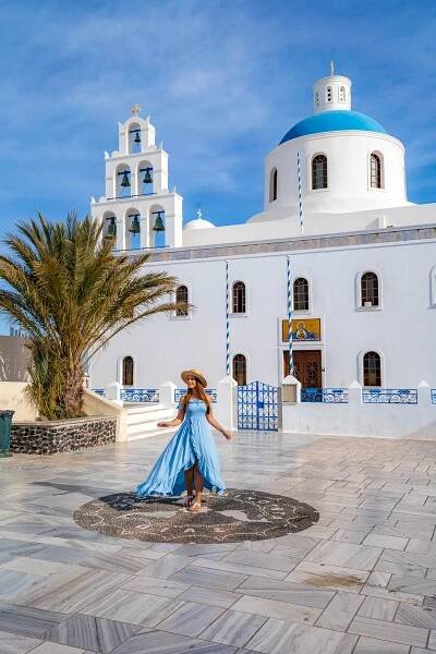 A woman in a flowing blue dress walking in front of the Church of Panagia Akathistos Hymn, showcasing the church's signature white facade, blue dome, and bell tower under a bright, sunny sky.