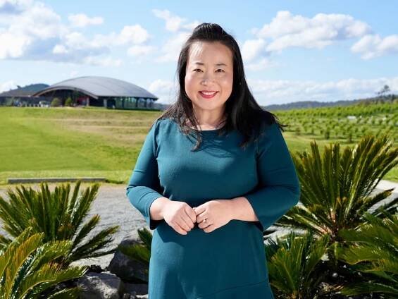 A woman with long black hair in a teal dress stands smiling with a modern building on a green hill in the background.