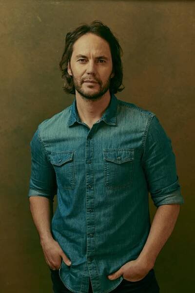 A man with dark hair and a beard in a denim shirt stands with hands in his jean pockets.