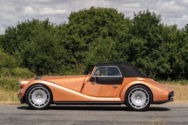 Klassisches Auto mit handgefertigter Karosserie und schwarzem Verdeck auf einer Strasse vor Bäumen.