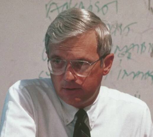 Fuller in a white button up and tie, wearing glasses and sitting in front of a dry erase board