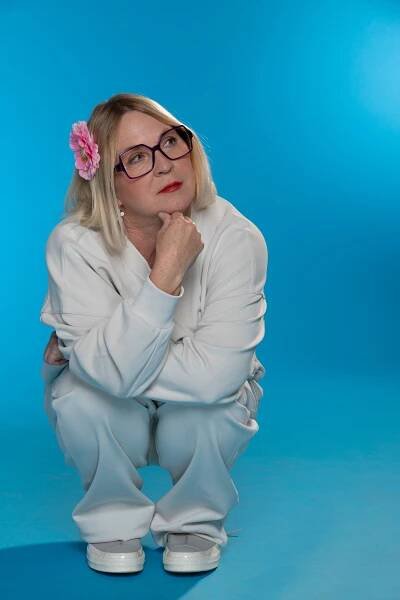 Carolyn Lawrence wearing glasses, wearing a casual white outfit with a pink flower in their hair, squats pensively against a blue background