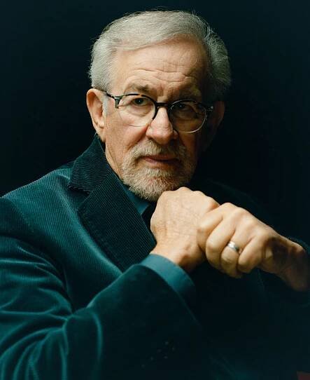 Steven Spielberg, wearing a green corduroy jacket, has his clenched his fists in front of him, one on top of the other as if holding a bat.