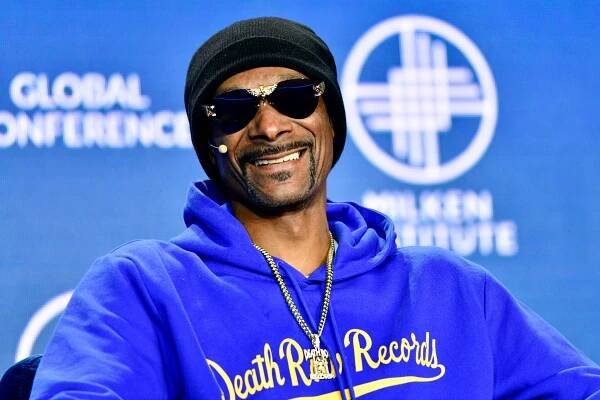 Snoop Dogg wearing sunglasses, a microphone headset, a beanie, and a blue hoodie with "Death Row Records" logo, smiles at a conference