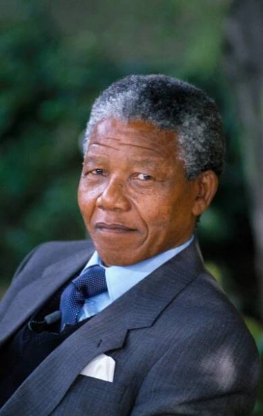 Elderly man in a suit and tie sits outdoors, looking thoughtful. Lush greenery is in the background