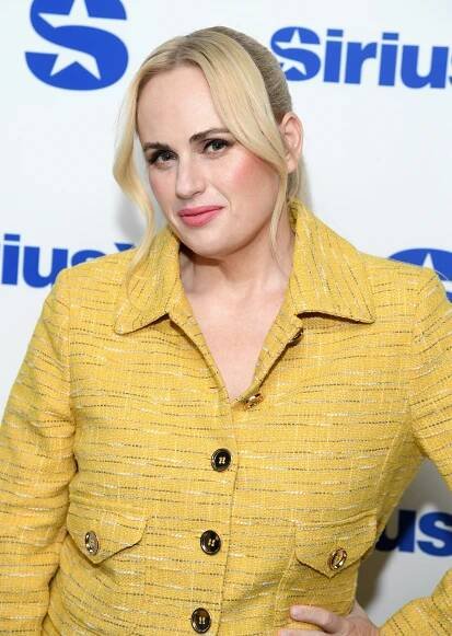 Rebel Wilson in a textured jacket poses at a media event backdrop