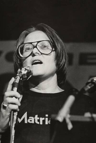 A black-and-white close-up of Barbara Love, in large glasses, speaking into a microphone.