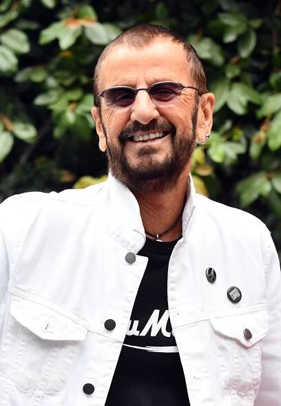 Rngo Starr wearing sunglasses and a white denim jacket smiling with green leaves in the background