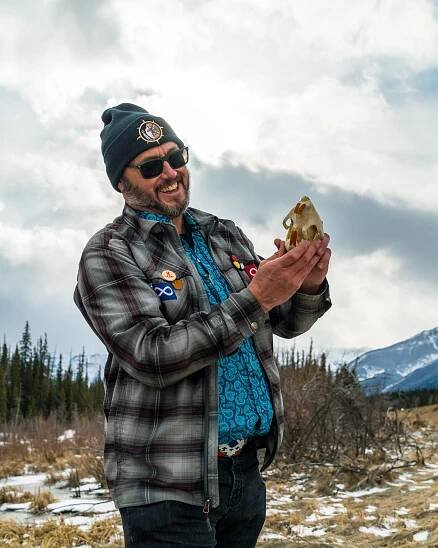 Man in outdoor gear, beanie, and sunglasses holds animal skull on snowy mountain landscape.
