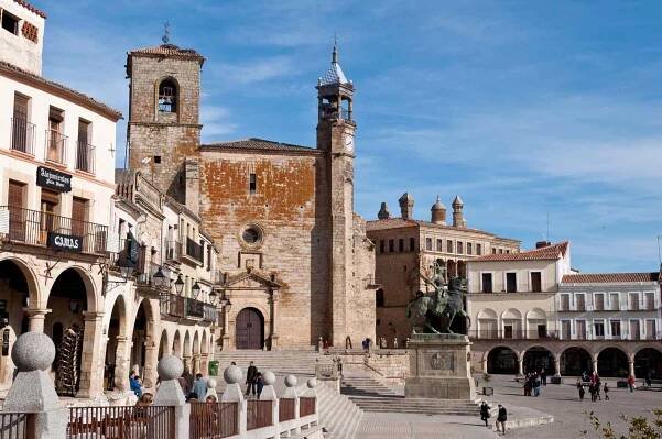 Historic town square featuring a statue and church, surrounded by buildings with arches and balconies