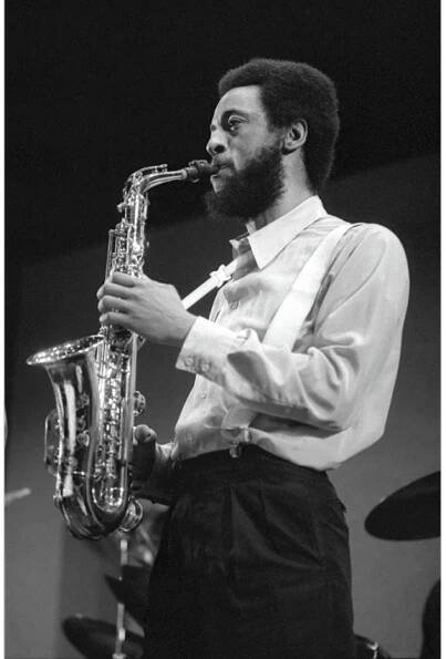Henry Threadgill playing a saxophone. He is bearded and wears black pants, a white shirt, and white suspenders.
