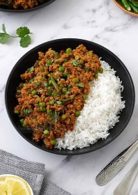 A black bowl containing white basmati rice and mince meat with green peas curry. Green chillies and curry leaves on the top. 