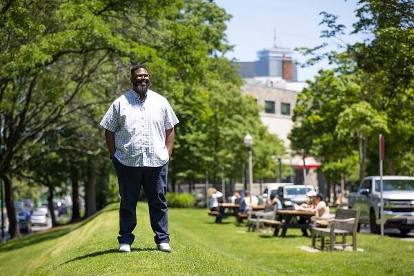 Photo: Anthony (Tony) Jack, a Black man wearing glasses, a light plaid collared shirt, and jeans, stands on a small grassy hill on a sunny day. Picnic tables filled with people can been in the blurry background behind him.
