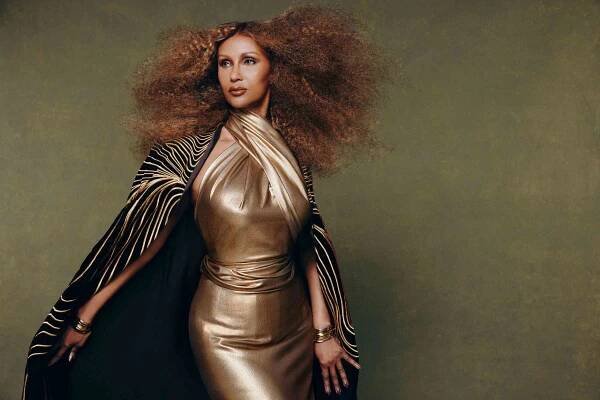 Iman in a gold halter dress with a patterned cape and curly hair.