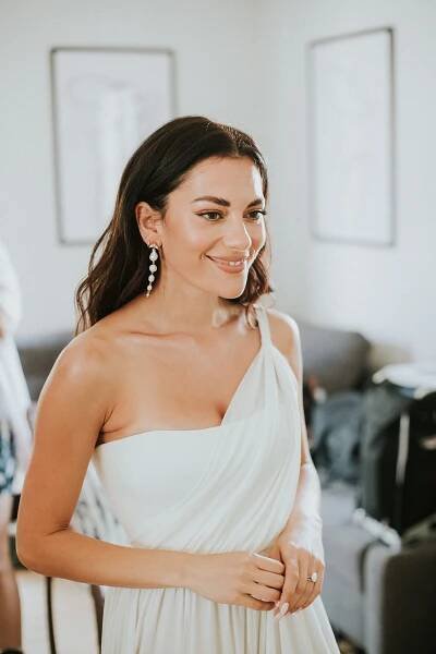 Bride Inbar Lavi smiling wearing a white one-shoulder wedding dress, chandelier earrings, and makeup