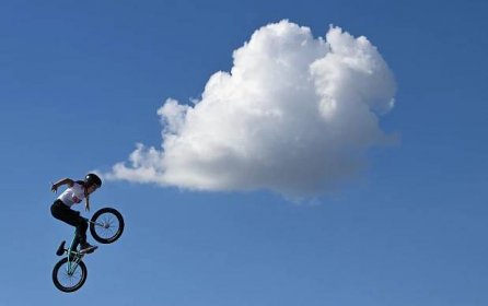 A competitor makes a jump with her BMX bike. A single large cloud floats in the blue sky beyond.