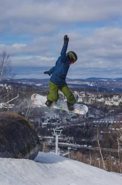 Une personne en planche à neige exécute un saut sur une pente de montagne enneigée. Un paysage hivernal avec des arbres, des télésièges et des collines lointaines est visible en arrière-plan.  
