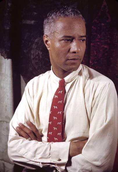 Harold Jackman, a British-born teacher, model and patron of the arts, looks to the side with crossed arms in a 1942 portrait. He wears a crisp, light-colored shirt with cuff links and a red print tie. 