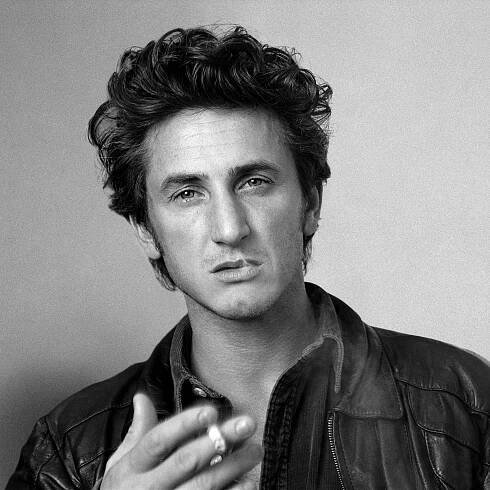 a black and white photo of a man in leather jacket holding his hand up to his face
