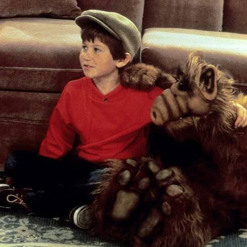 Benji Gregory, a young boy wearing a cap, sits in front of a couch with the puppet known as ALF. Their arms are wrapped around each other. 