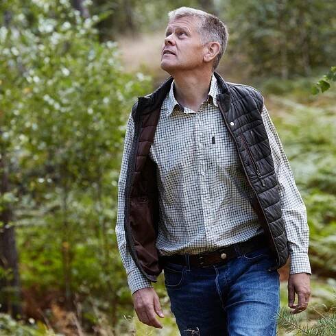 A man in a checkered shirt and a vest walking in the forest.