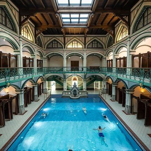 Das Bild zeigt ein großes Hallenbad in einem alten, schönen Gebäude. Das Becken ist rechteckig, das Wasser ist hellblau. Einige Menschen schwimmen darin. Um das Becken herum stehen kleine Umkleidekabinen mit braunen Vorhängen. Oben gibt es einen Balkon mit grünen Geländern. Das Dach besteht aus Holz und Glas, durch das Licht hereinkommt. Foto: Philipp Herfort Photography