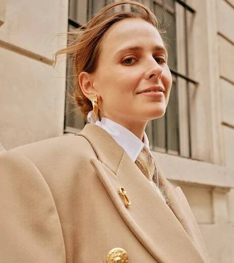 A woman in a stylish beige oversized blazer and white shirt poses confidently outdoors, adorned with...