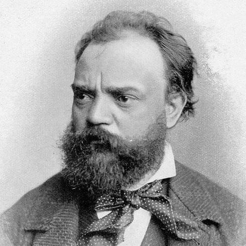 A black-and-white vintage image of Czech composer Antonin Dvorak. He is a white man with a thick beard and dark hair. He wears a dark jacket with a light stripe pattern, a matching vest, and a white dress shirt. He also wears a polka-dotted cravat or scarf tied into a bow underneath his dress shirt’s collar.