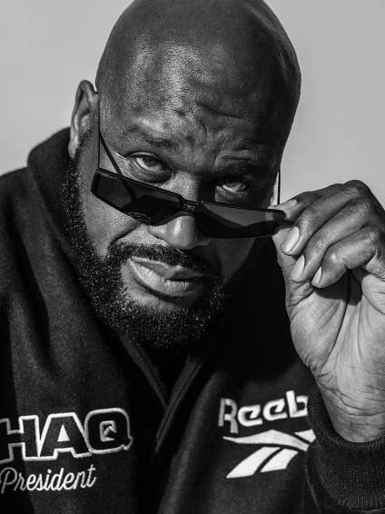 A large bald man with a beard looks at the camera while pulling his sunglasses down. His jacket says “Shaq President” and Reebok.