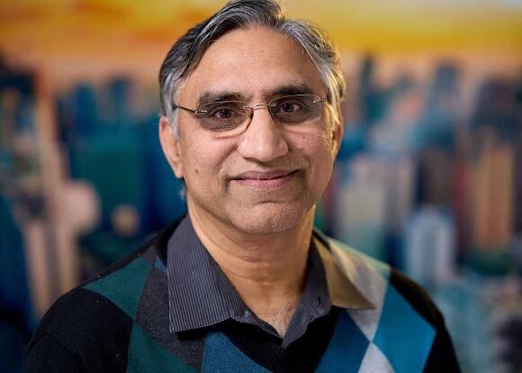 Professional portrait of a person in dark sweater against cityscape backdrop.