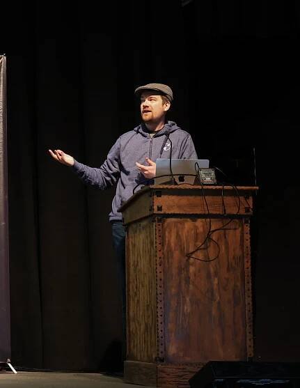 Alex Anderson on stage gesturing at the screen wearing a blue hoodie