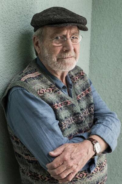 A bearded man wearing glasses and a cap dressed in a blue shirt and sweater vest looks at the camera. 