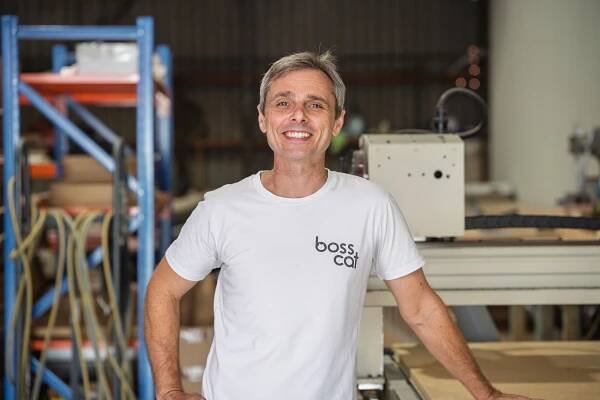 Person in white t-shirt with logo smiling in a workshop or manufacturing space with shelving in the background.