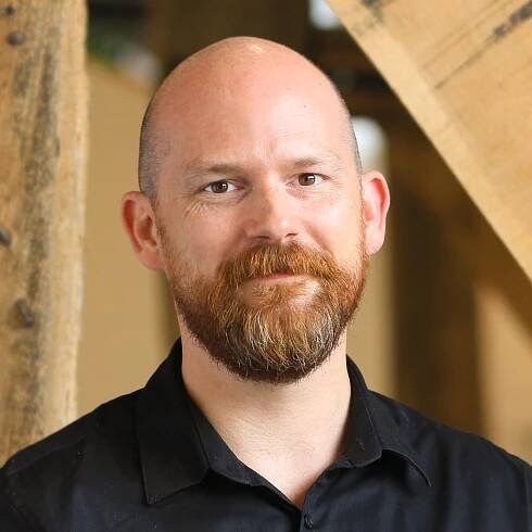 Headshot of a consultant with a ginger beard, wearing a black polo shirt. He has a serious expression.