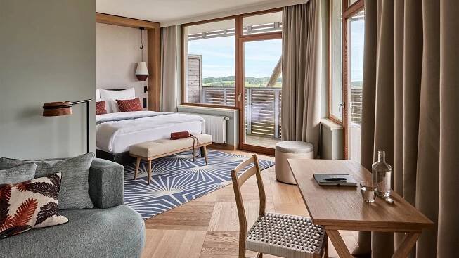 Modernes Hotelzimmer mit Bett, Sitzecke, Tisch und Balkon mit Landschaftsblick.