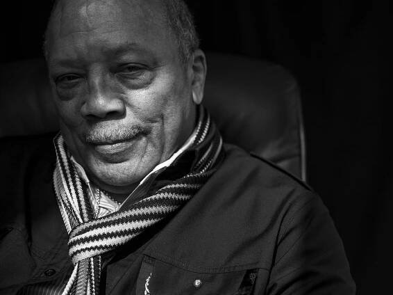 A black-and-white image of Quincy Jones wearing a scarf and a jacket, with a small mustache.