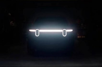 Teaser image for the Rivian R2 EV. Two vertically oval headlights along a sleek horizontal light bar hint at the front of the vehicle. Otherwise nearly black image of a garage.