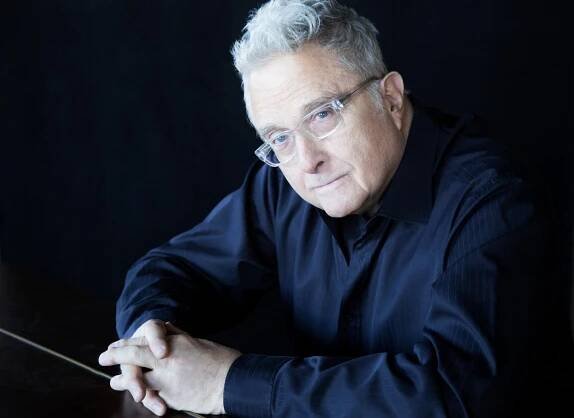 An older man with gray hair and glasses sits at a table, wearing a dark shirt and looking towards the camera, reminiscent of Randy Newman contemplating his renowned music catalog.