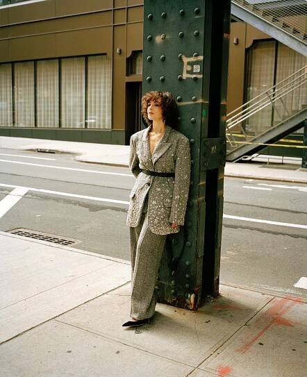 A woman in a black-and-white herringbone-patterned pantsuit, with a black belt, is leaning with her back against a green-painted steel column on an empty city street.