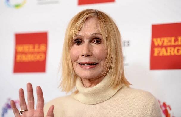 Sally Kellerman waving at the camera in a cream turtleneck. 