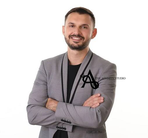 Professional Headshot of someone in a gray blazer with black trim and black shirt standing with arms crossed against white background.
