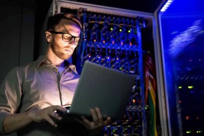 Seriózní zaměstnaný mladý inženýr v brýlích, které stojí proti síťovému serveru s planoucím senzorem v tmavé místnosti a používá přenosný počítač při sledování síťových systémů — Stock obrázek