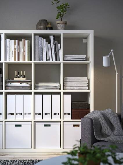 Ein weißes KALLAX Regal 4x4 mit Zeitschriftensammlern, Büchern und weißen Papierboxen steht an einer Wohnzimmerwand. 