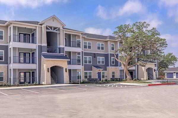 A modern multi-story apartment building with a light gray exterior, featuring balconies and stone accents. In front, there is a spacious parking lot with empty parking spaces. A large tree provides shade next to the building, under a clear blue sky.