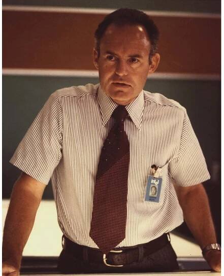 A man wearing a striped shirt and tie leans on a desk. He has a badge clipped to his shirt and appears to be in an office or control room setting, with a focused expression. The background is blurred.