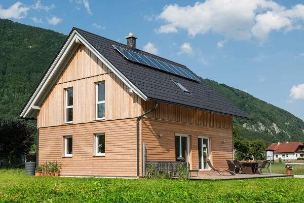Modernes Einfamilienhaus in Österreich mit Holzverkleidung und Solarpanelen auf dem Dach, im Hintergrund grüne Berge unter blauem Himmel.