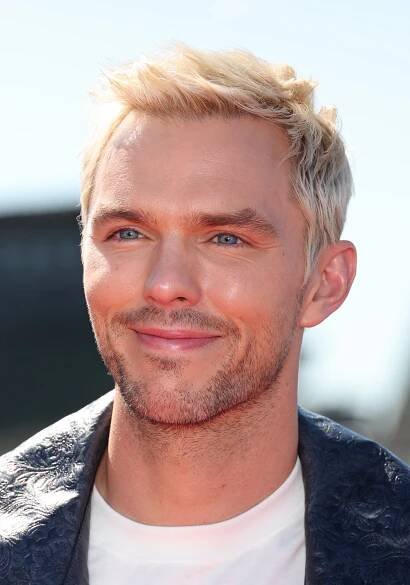 Nicholas Hoult with short hair and light stubble smiles at a public event, wearing a patterned jacket over a plain shirt