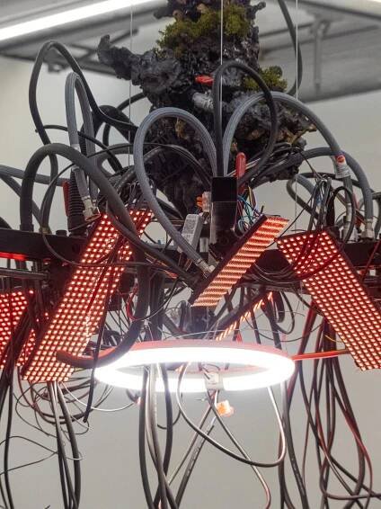 An abstract installation featuring tangled wires, red LED lights, and a central illuminated ring, with a moss-covered structure above.
