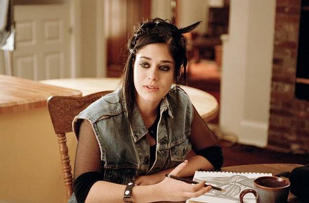 Lizzy Caplan wearing a denim vest over a black top, sitting at a table with a sketchbook and coffee mug in "Mean Girls"