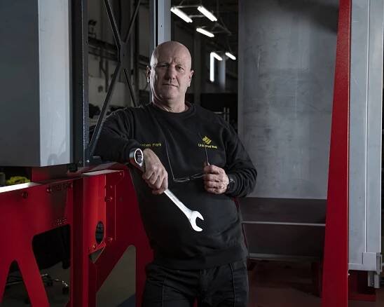 Man in branded Universal Wolf sweatshirt holding a wrench and leaning against red metalwork